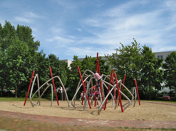 Klettergerät aus roten und silbernen Metallbögen auf Sand vor Laubbäumen