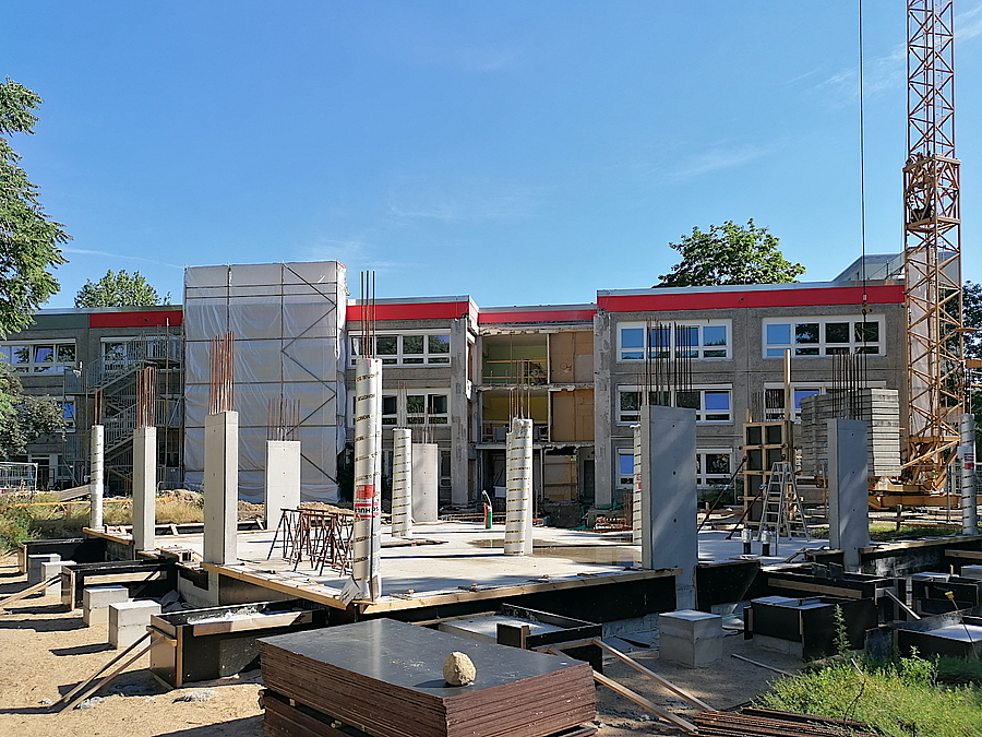 Baustelle mit Betonstützen, dahinter Bestandsgebäude der Kita