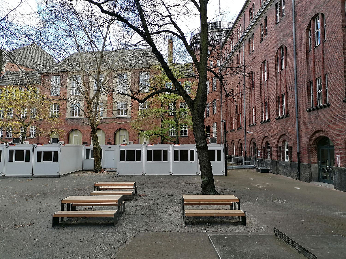 Schulhof mit Tisch-Bank-Kombinationen und Containern, Backsteingebäude, Bäume im Frühjahr