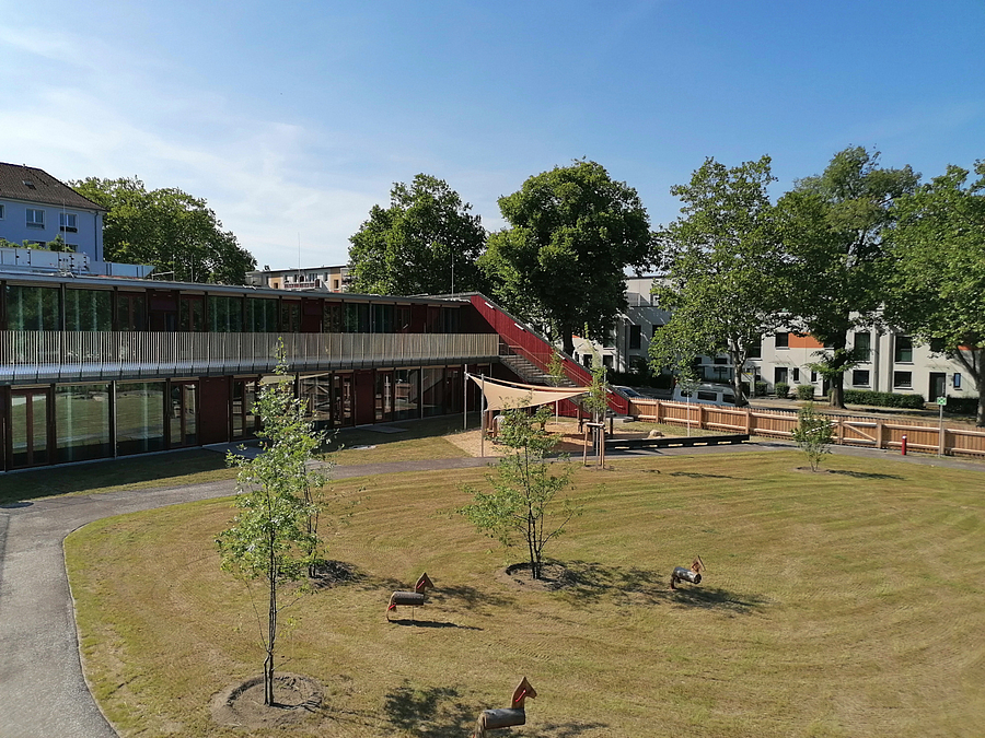 Kitagarten mit Jungbaeumen und Holzpferden, zweistöckiges Gebäude mit Terrasse und Freitreppe als Begrenzung