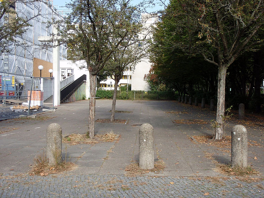 Platz mit Pollern, Bäumen und eingerüsteter FAssade, hohes christliches Kreuz aus Beton