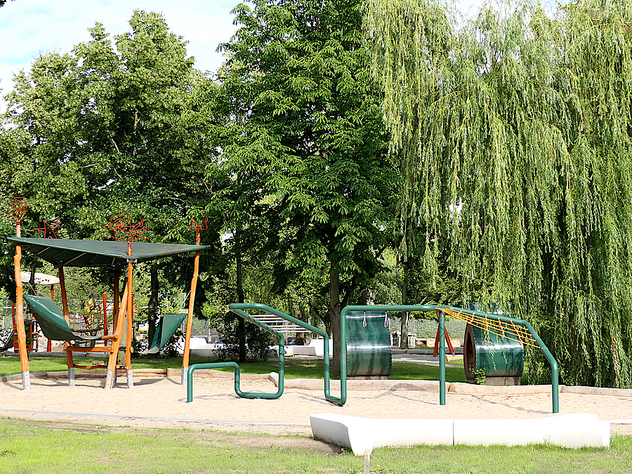 Kletter- und Hangelparcours aus Stahl und Netz auf Sand, dahinter überdachter Pavillon mit Hängematten, alte Bäume