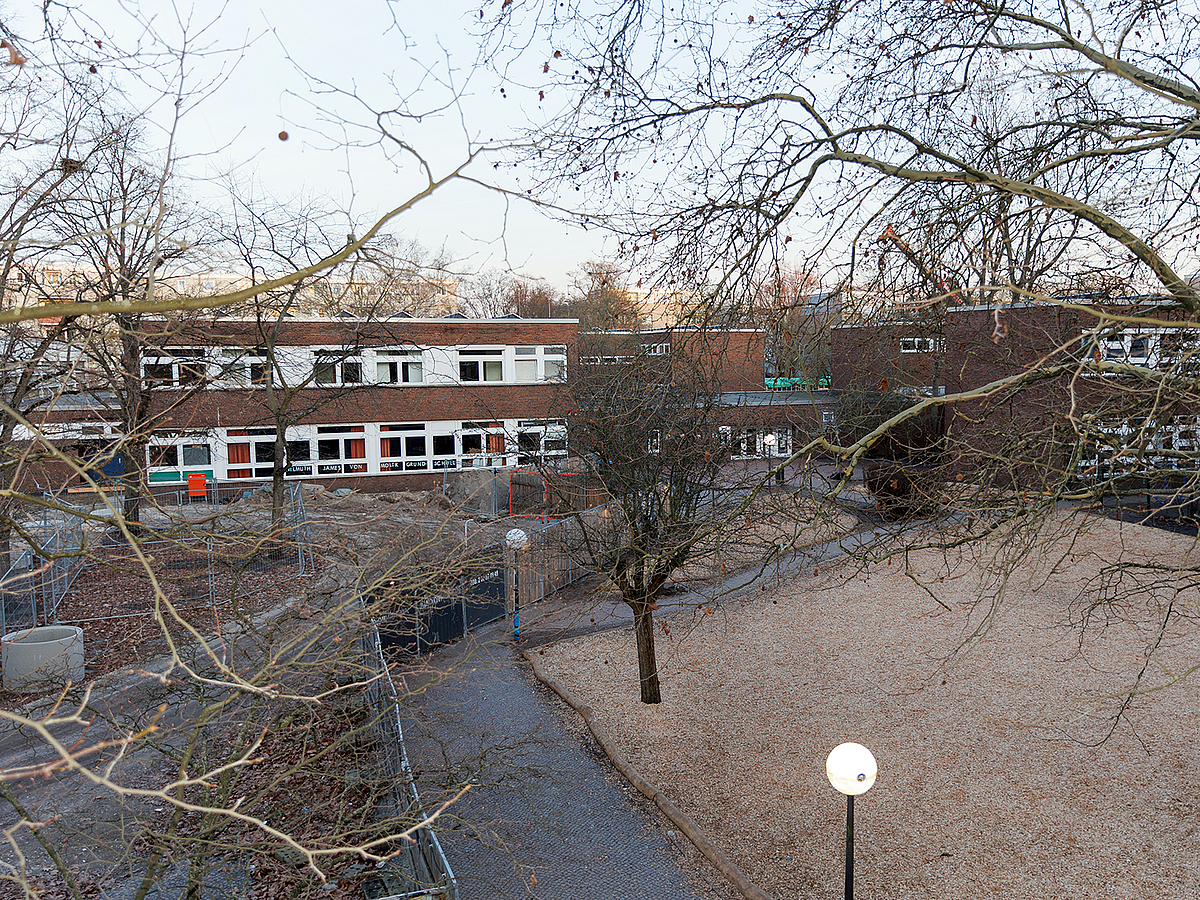 Blick auf leere Fläche mit Holzhäckseln, winterlich kahle Bäume und 2-stöckiges Schulgebäude