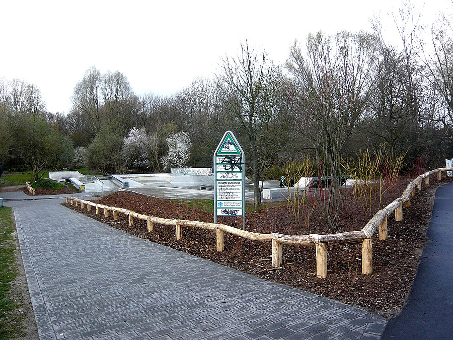 Weg vor neuer Pflanzfläche mit Gehölzen, Schild Geschützte Grünanlage, dahinter Skatefläche