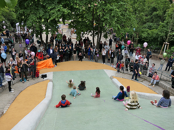 Blick von oben auf grüne Hangrutsche mit Kindern und Publikum am Auslauf