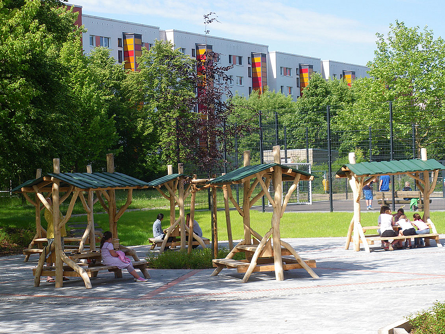 Freisitz mit überdachten Sitzmöbeln Überdachte Holzbänke auf Pflasterfläche, dahinter bunte Plattenbauten, Bäume