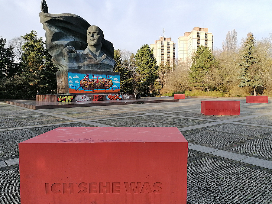 Platz mit großen, roten Quadern. Eine Aufschrift "Ich sehe was", dahinter Denkmal, Hochhäuser