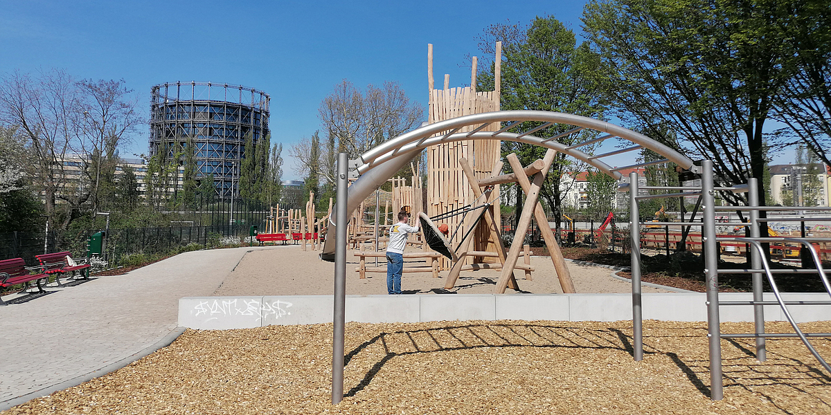 Hangelbrücke auf Spielplatz mit hölzernem Kletterturm, im Hintergrund Gasometer