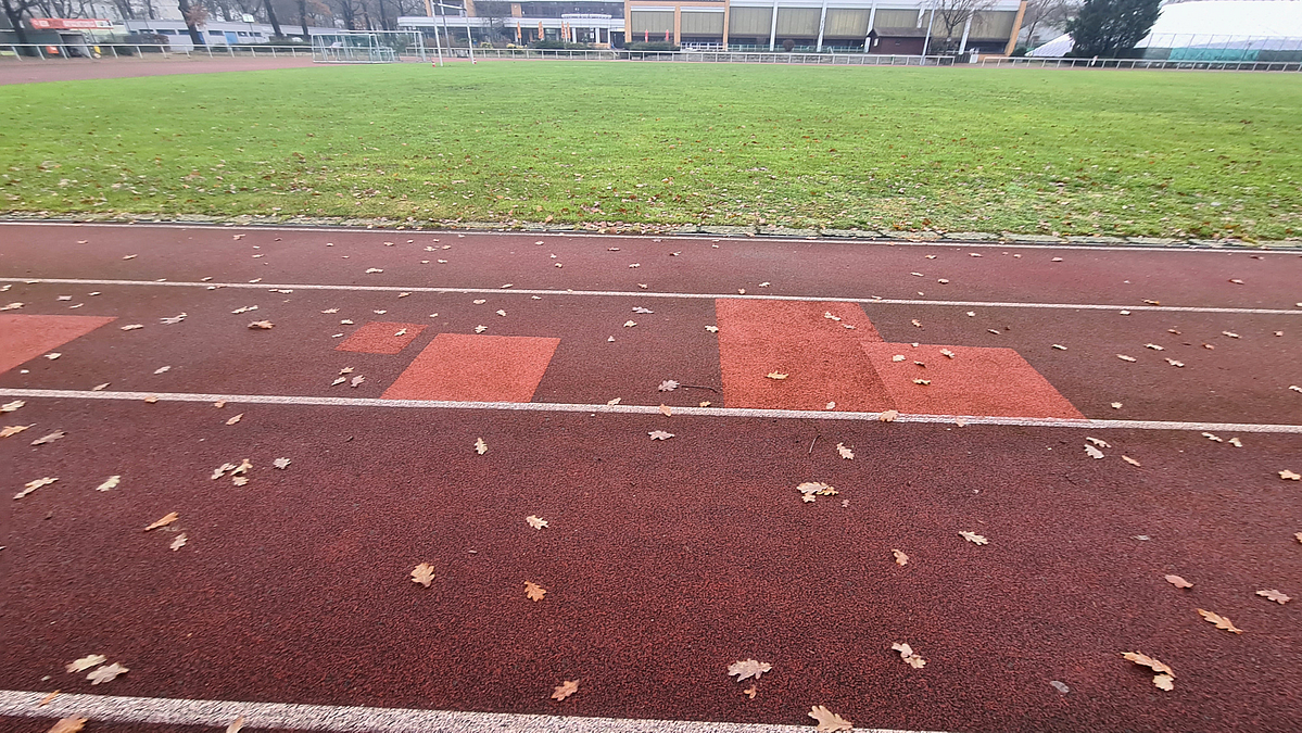 Geflickte Kunststoff-Laufbahn vor Sportplatz-Rasen