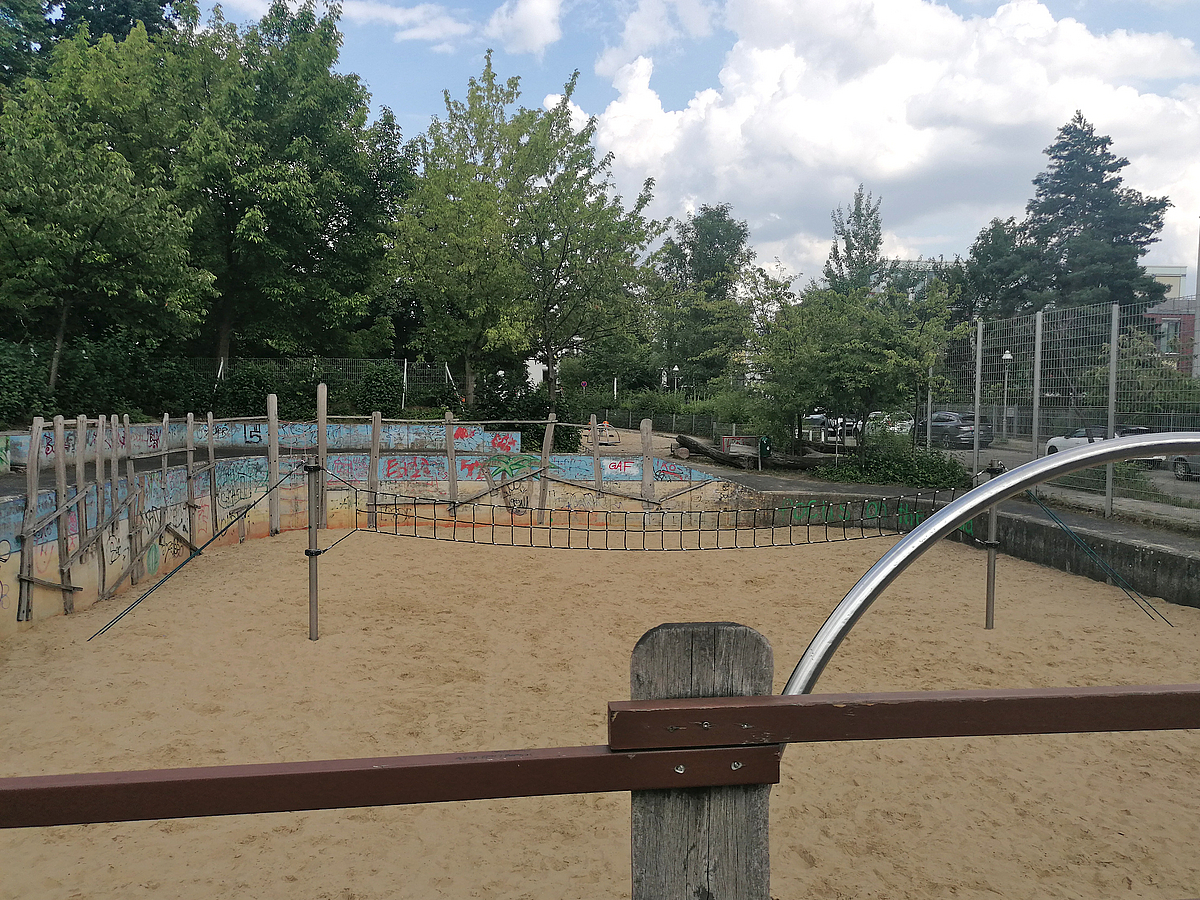 Sandfläche mit Beachvolleyballnetz und Holzbalken an einer blauen Stützwand mit Graffiti, Laubbäume im Hintergrund
