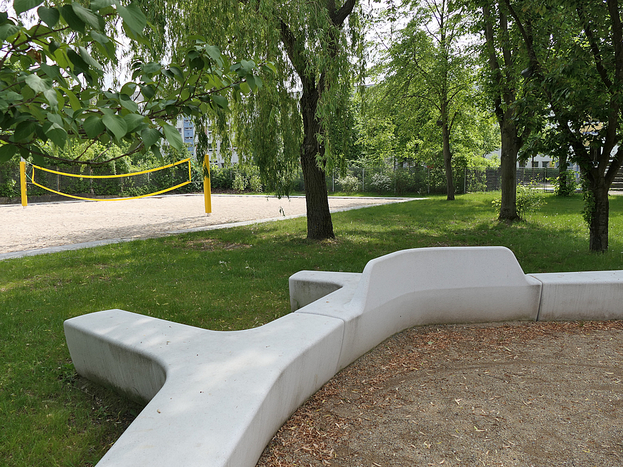 Organisch geformtes Betonsitzelement, dahinter Rasen mit Bäumen, Beachballfeld mit Netz