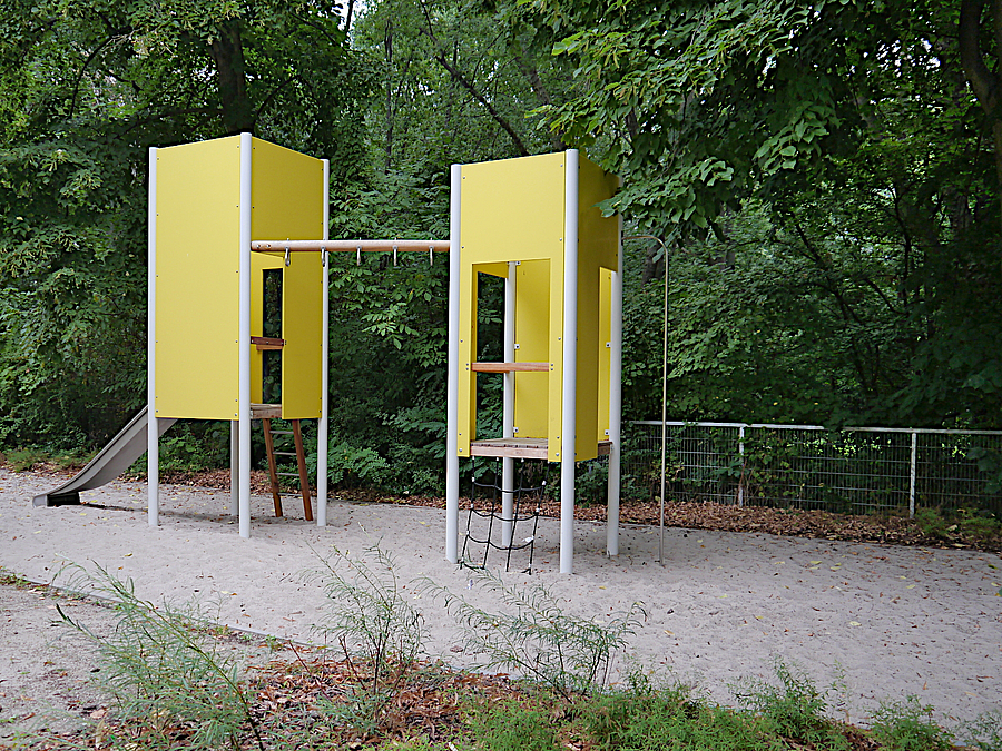 2 schmale gelbe Spielhäuser auf Stelzen im Sand, dahinter Grün