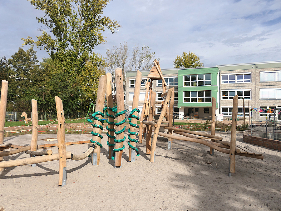 Kita-Typenbau mit grünem Akzent, davor Sandspielfläche mit Balancierlandschaft aus Holz mit grünen Seilen