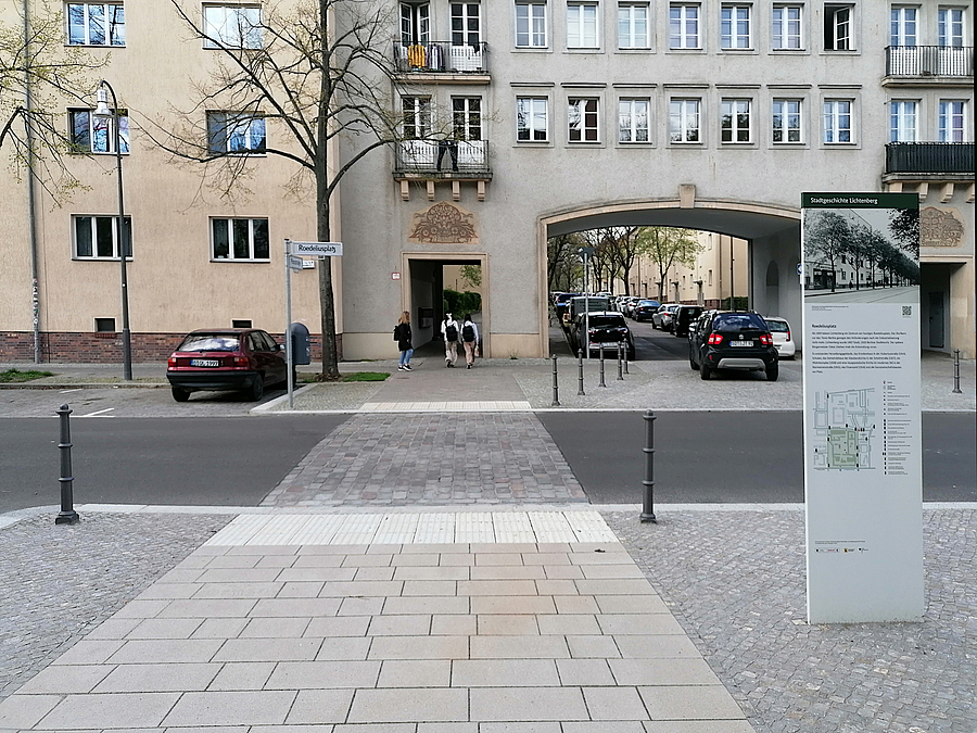 Plattenweg auf gepflastertem Platz setzt sich über die Straße fort, Poller, Infotafel zur Geschichte