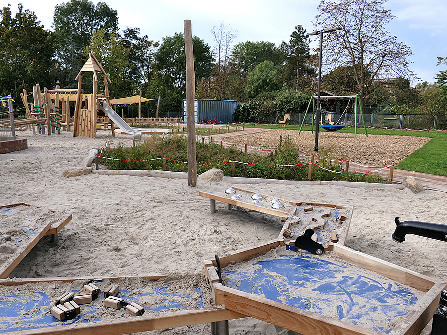 Sandflaeche mit Matschanlage und Rutschenturm aus Holz, Rasen, Nestschaukel