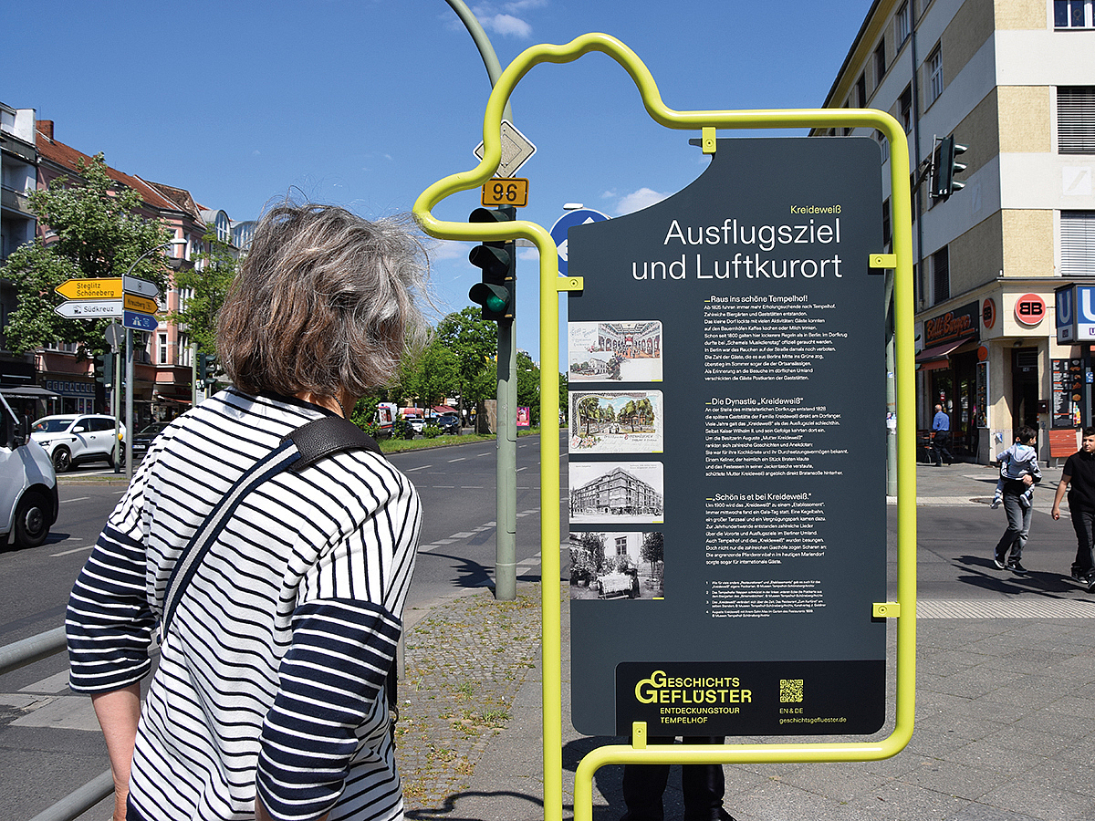 Frau betracthet Infostele auf der Straße mit gelbem Metallrahmen in Form eines Hutes und schwarzer Tafel zum Thema Ausflugs-Lokal