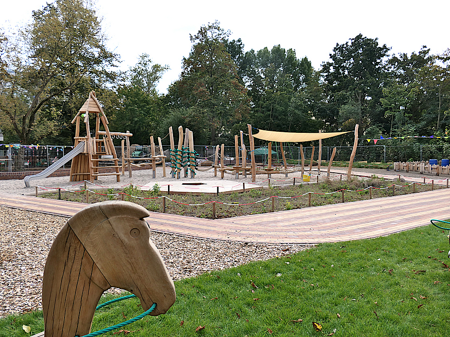 Kitagarten Sandflaeche mit Balancierstrecke und Rutschenturm aus Holz und Seilen mit Sonnensegel, Beet, Rundweg, Rasen, im Vordergrund Pferdekopf auf Holz