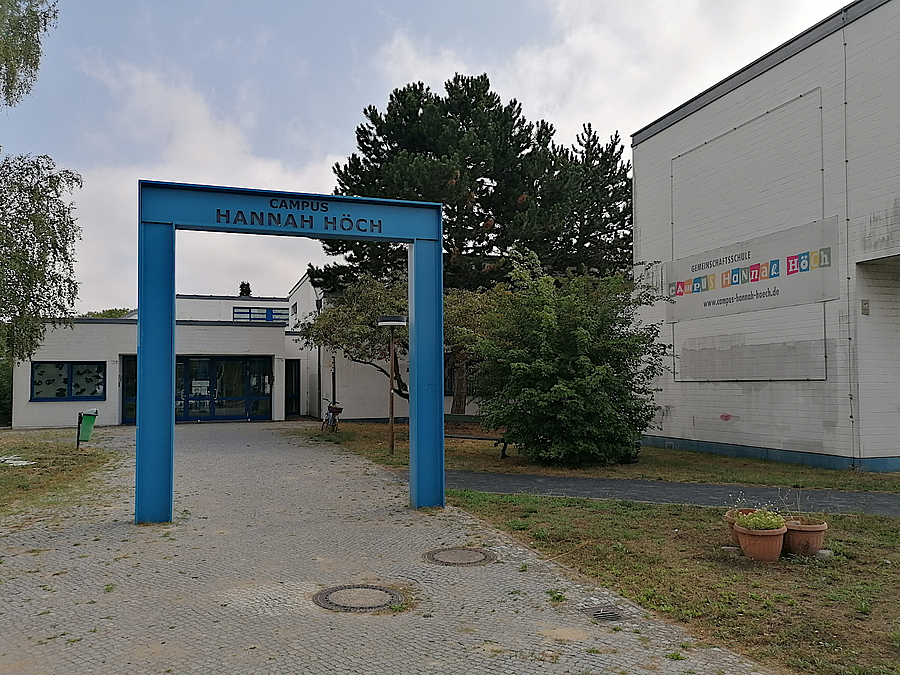 Der Eingangsbereich der Oberschule wird neu gestaltet Großer, blauer Rahmen auf Weg vor flachem, hellem Gebäude, Bäume