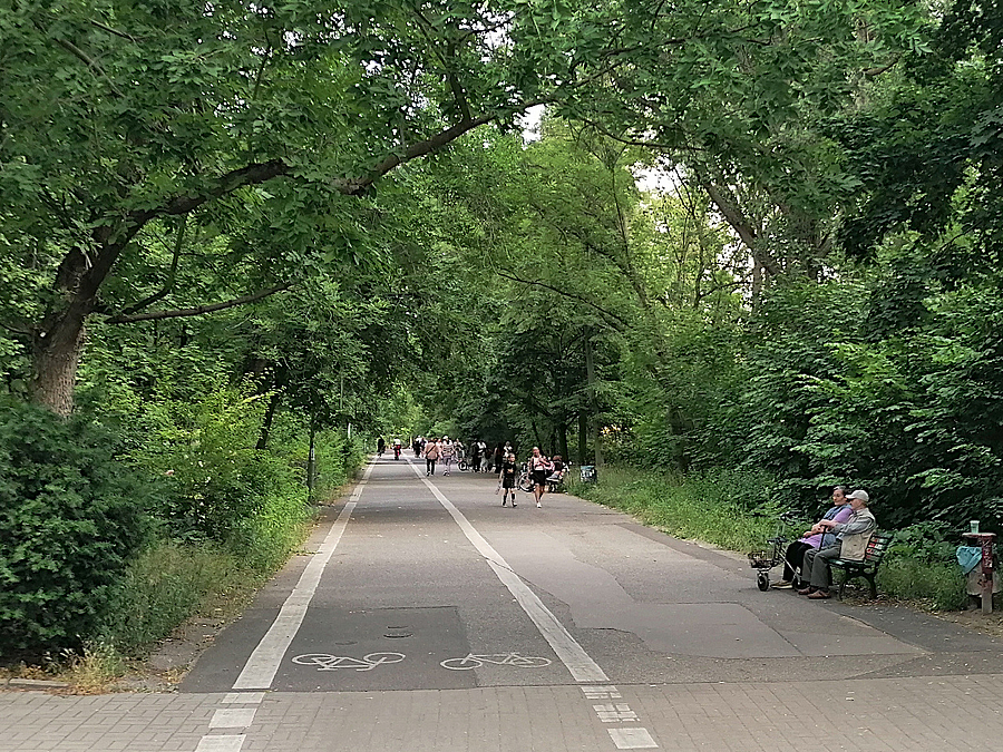 Asphaltweg mit markiertem Fahrradstreifen, links und rechts Grün. Älteres Paar auf der Bank, im Hintergrund weitere Menschen