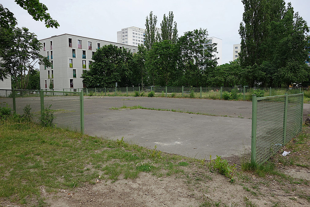 Große Flächen sind versiegelt und kaum nutzbar Umzäunter Asphaltplatz mit Unkraut, dahinter Neubau