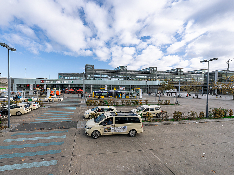 VErsiegelter Platz mit Taxis und Bussen, aufgemalter Gehweg aus blauen Zebrastreifen, im Hintergrund Bahnhof