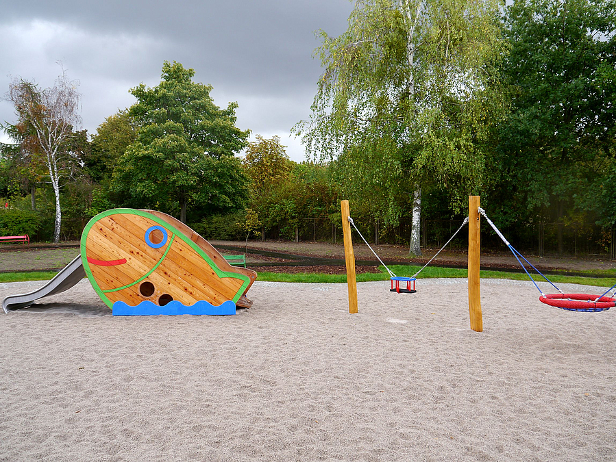 Sandfläche mit 2 Kleinkindschaukeln und Kleinkindrutsche mit Seitenwand als bunter Fisch