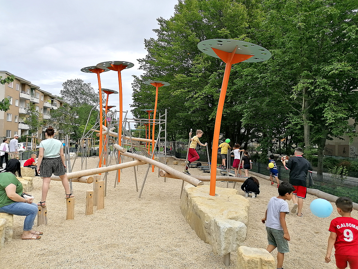 Spielplatz auf Sand mit Sandsteinen, Kletterbalken, gebogenen, orangefarbenen Pfeilern mit grünen Tellern als Abschluss, Kinder u. Erwachsene