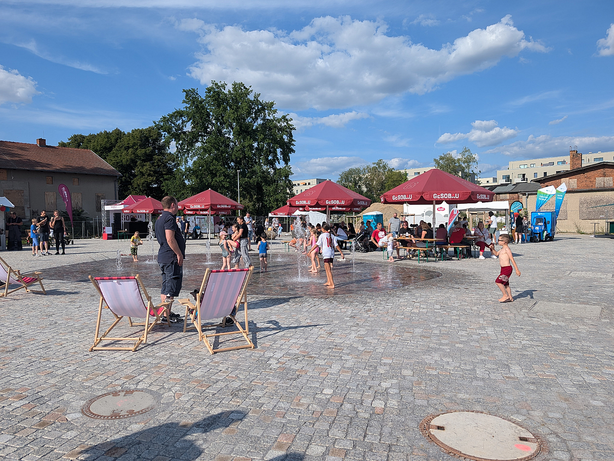Belebter Platz mit Wasserspiel, Sonnenschirmen und Liegestühlen
