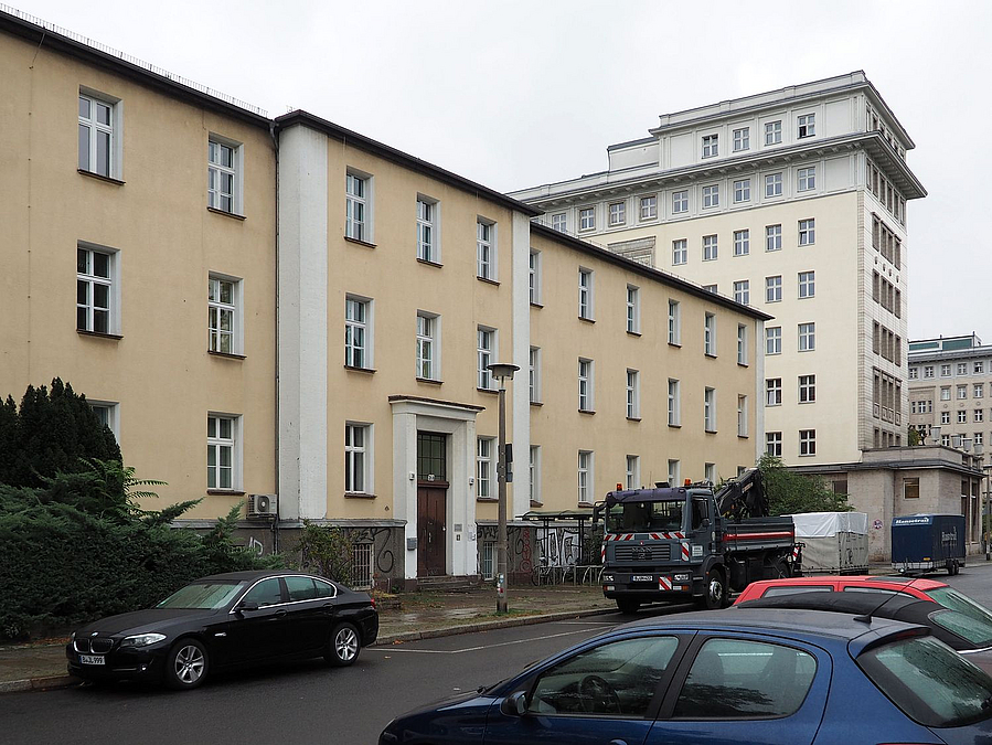 Heller dreistöckiger Gebäuderiegel aus der Nachkriegszeit an einer Straße, dahinter höherer Querriegel
