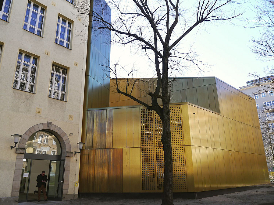 Schulgebäude mit Tor im Anschnitt und messingfarbener unregelmäßiger Baukörper