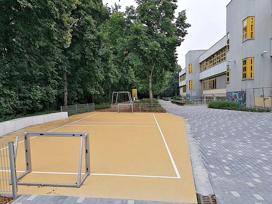 Bolzplatz mit Minitoren und hellem Belag, Weg, Schulgebäude, Laubbäume