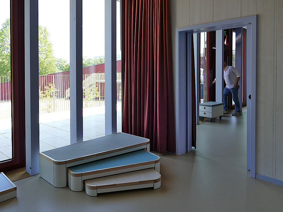Blick durch bodentiefes Fenster auf Terrasse, roter Vorhang, zusammenschiebbare Podeste als Treppe für Kinder, Tür mit violettem Rahmen, helle Wand