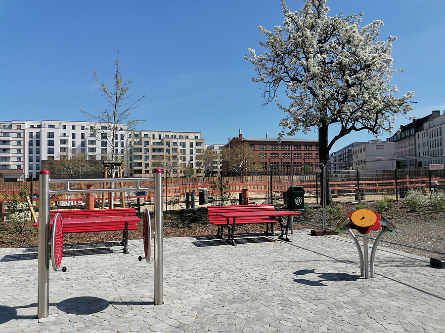 Rote Bänke und Tisch, Fitnessgerät für Schultern, stilisierte Blumen auf glatter Pflasterfläche, blühender Obstbaum