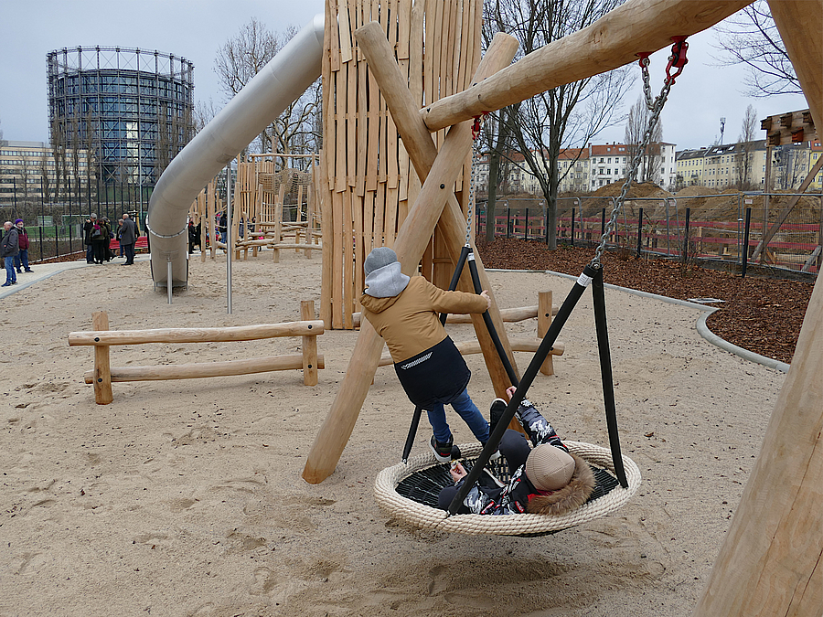Spielplatz mit Nestschaukel in Nutzung und großer Tunnelrutsche, im Hintergrund Gasometer