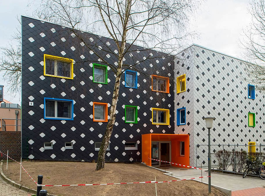 Mit schwarzen bzw. weißen Schieferplatten verkleidete Fassade mit Muster und farbigen Fensterbrüstungen, davor abgezäunte Fläche mit winterlicher Birke, ohne Rasen, gepflasterter Weg zum Eingang