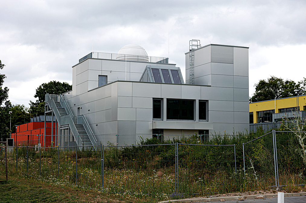 Betonbau mit großem Balkon, Turm und kleiner Kuppel und Außentreppe, davor bewachsene Baustelle