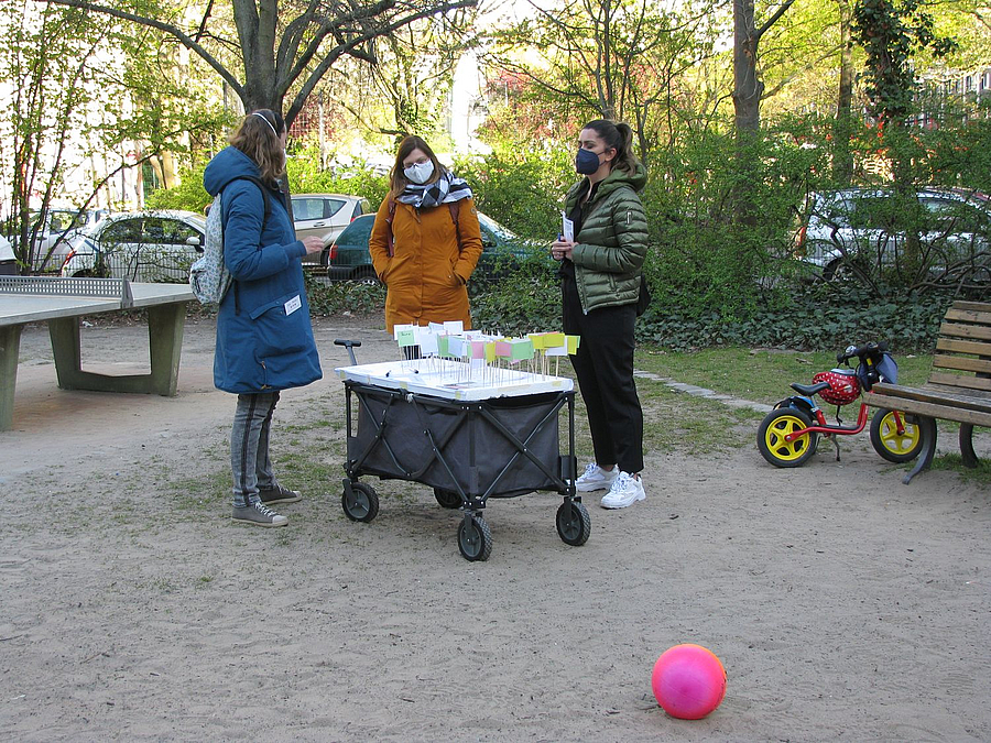Handwagen mit Plan und Fähnchen und 3 Personen auf Spielplatz