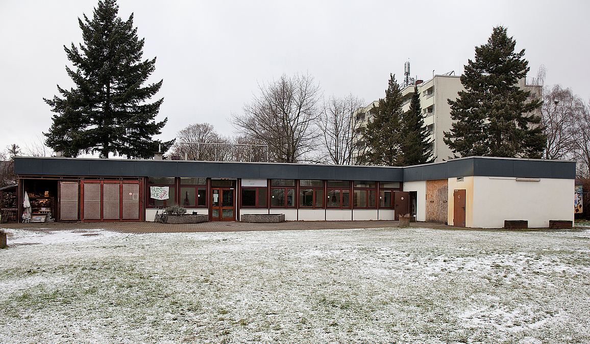 Zweiflügeliger Flachbau auf winterlicher Wiese, dahinter Plattenbau und Fichten