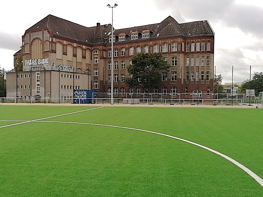 Grüner Fußballplatz, dahinter historisches zweiflügeliges Schulgebäude