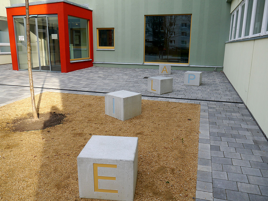 Betonwürfel, Holzhäckselfläche mit jungen Baum, roter Eingang, grüne Fassade