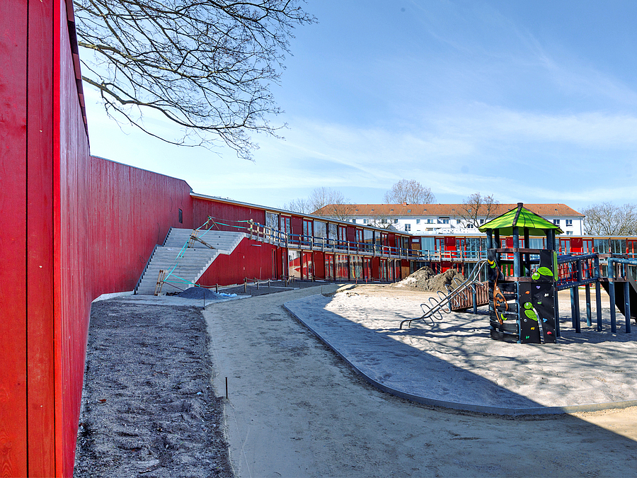 Blick an roter Holzwand vorbei auf sandigen Innenhof mit Spielgerät, Freitreppe am Gebäuderiegel