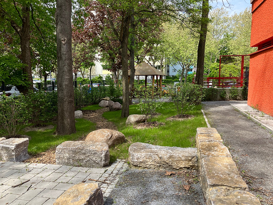 Gebäudeecke mit grünem Umfeld, Bäume, Findlinge, Granitquader, Holzpavillon