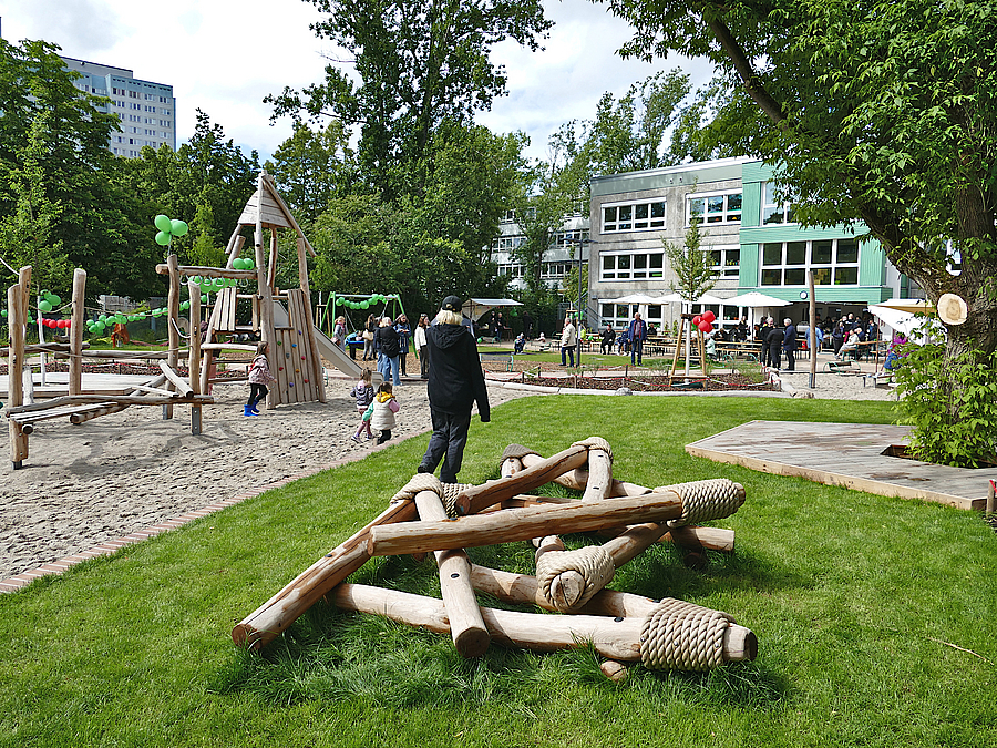 Balancierstämme auf Wiese, Sandspielfläche mit Kletterparcours, dahinter Kita-Typenbau mit grünen Akzenten