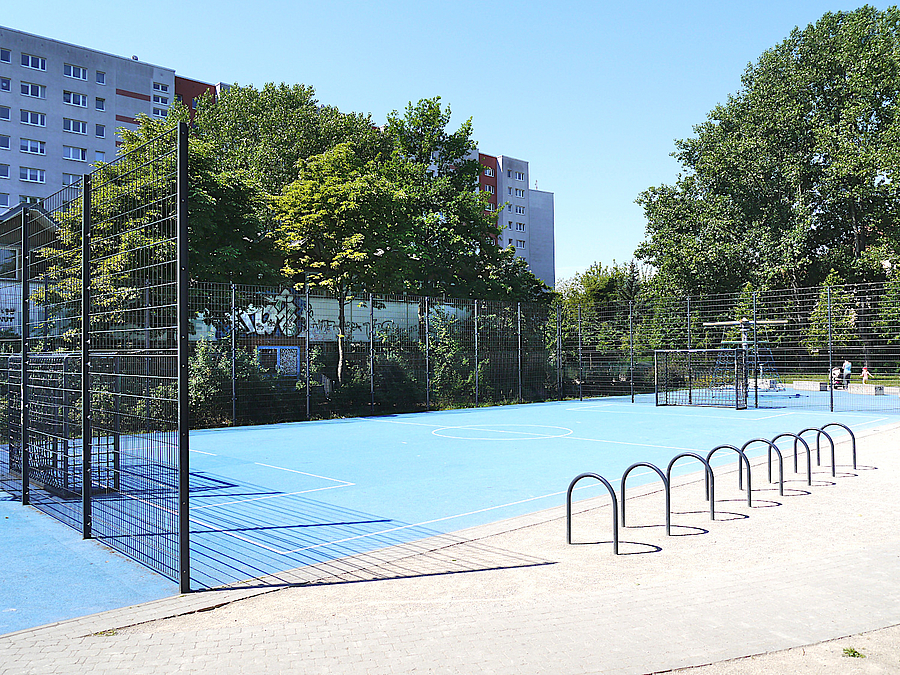 Bolzplatz mit blauem Boden, an einer Seite offen, dahinter Bäume und Elfgeschosser