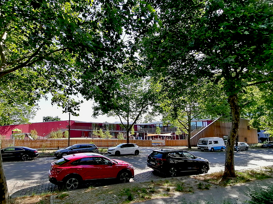 Zweistoeckigen Gebaeude in rot mit Fensterfront und Garten mit Sonnensegel, Holzlatten-Fassade zur Straße mit Bäumen und parkenden PKW