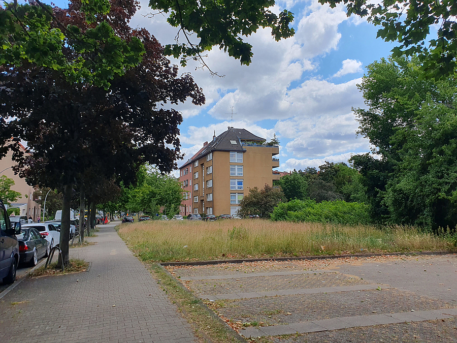 Parkplatzfläche und Wiese an der Straße, dahinter Mehrfamilienhaus