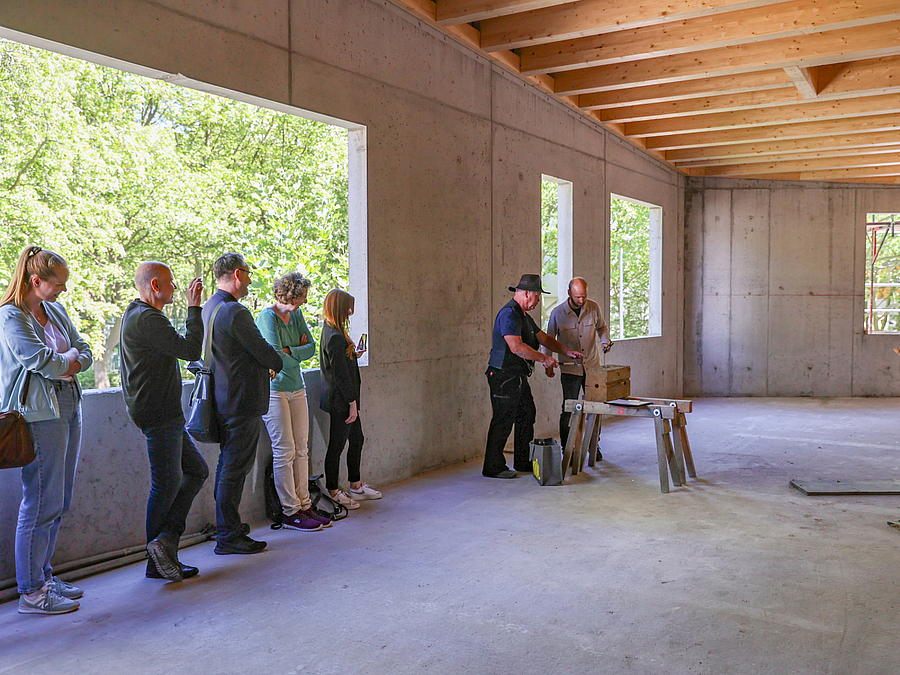 Menschen stehen im Betonrohbau mit Balkendecke mit großen Fensterlöchern