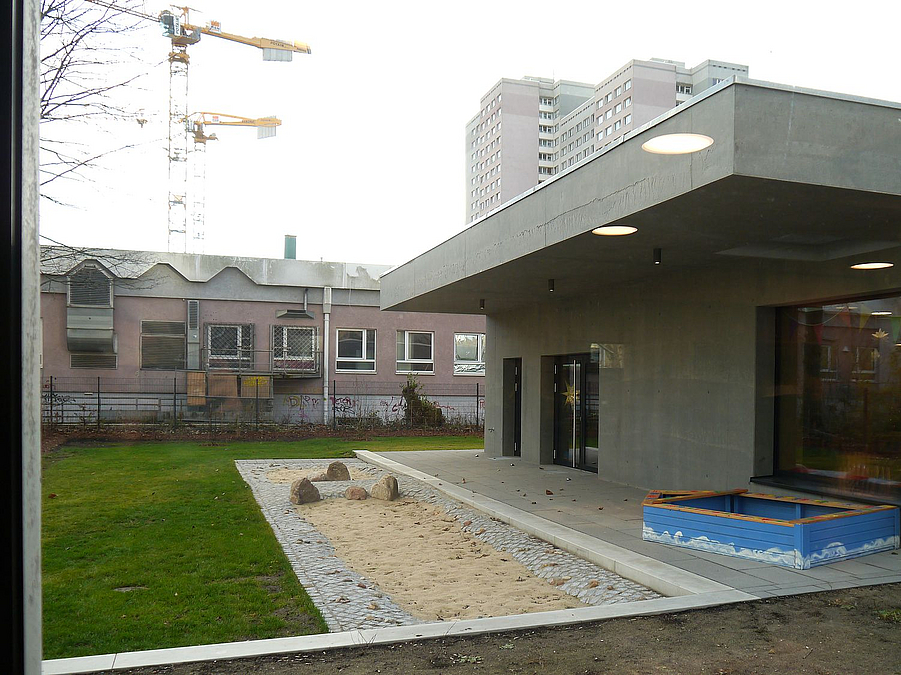 Sandfläche und Rasen vor Betonbungalow mit vorgezogenem Dach