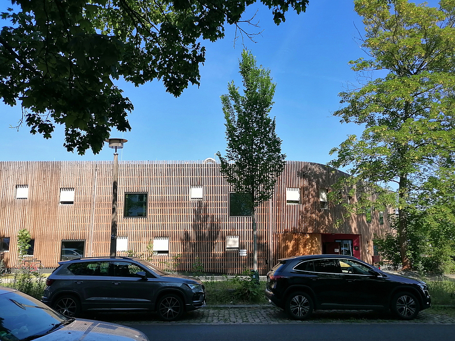 Holzlatten-Fassade eines zweistöckigen Gebäudes mit asymmetrischen Fenstern,  davor Straße mit Bäumen und parkenden PKW