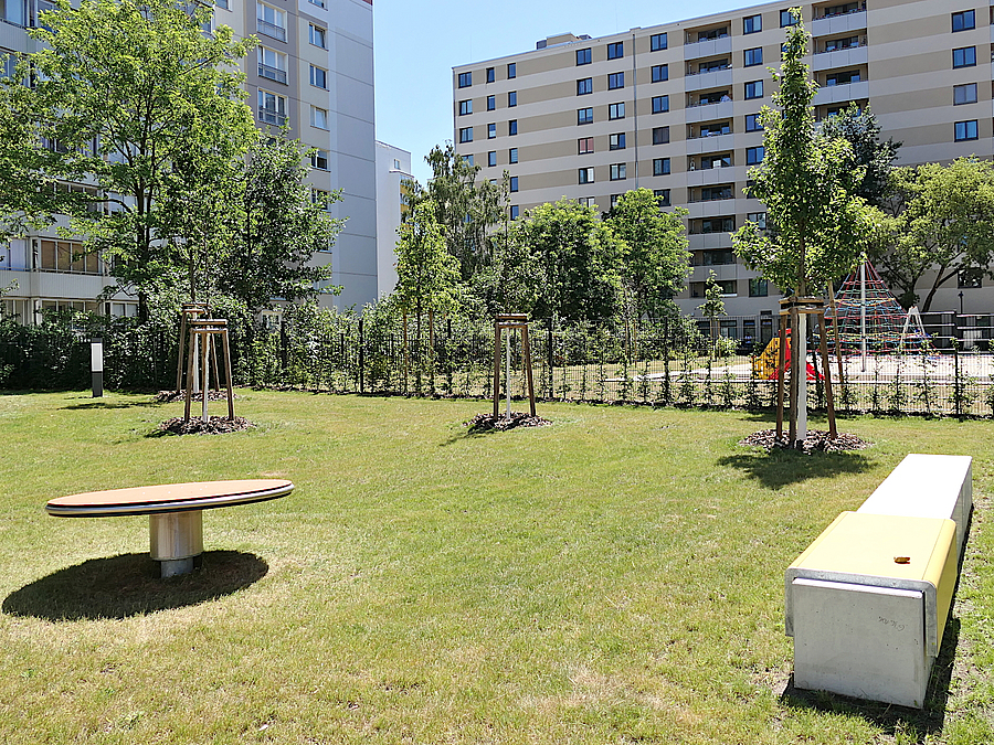 Garten mit Drehscheibe, Betonbank und jungen Bäumen, im Hintergrund moderne Wohnhochhäuser